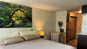 Une chambre avec un grand lit, une table de chevet avec une lampe, une petite table en bois ornée d’une plante et un tableau représentant un arbre accroché au mur.