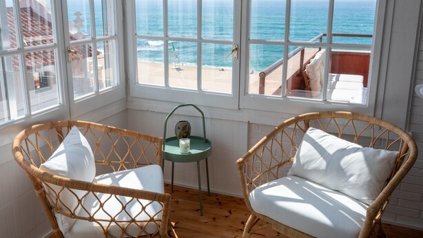 Une pièce avec des fauteuils en osier, une petite table sur laquelle est posée une bougie, et une vue sur l’océan par de grandes fenêtres.