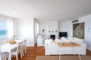 A modern living room with a white sofa, a dining table with chairs, a fireplace, and a large window overlooking the ocean. - Victoria - in front of the beach (Sant Vicenç de Montalt)