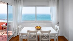 Un coin repas avec une table et des chaises, un saladier rempli de fruits et une vue sur l’océan par de grandes fenêtres.