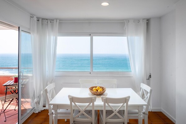A dining area with a table and chairs, a bowl of fruit, and a view of the ocean through large windows. - Victoria - in front of the beach (Sant Vicenç de Montalt)