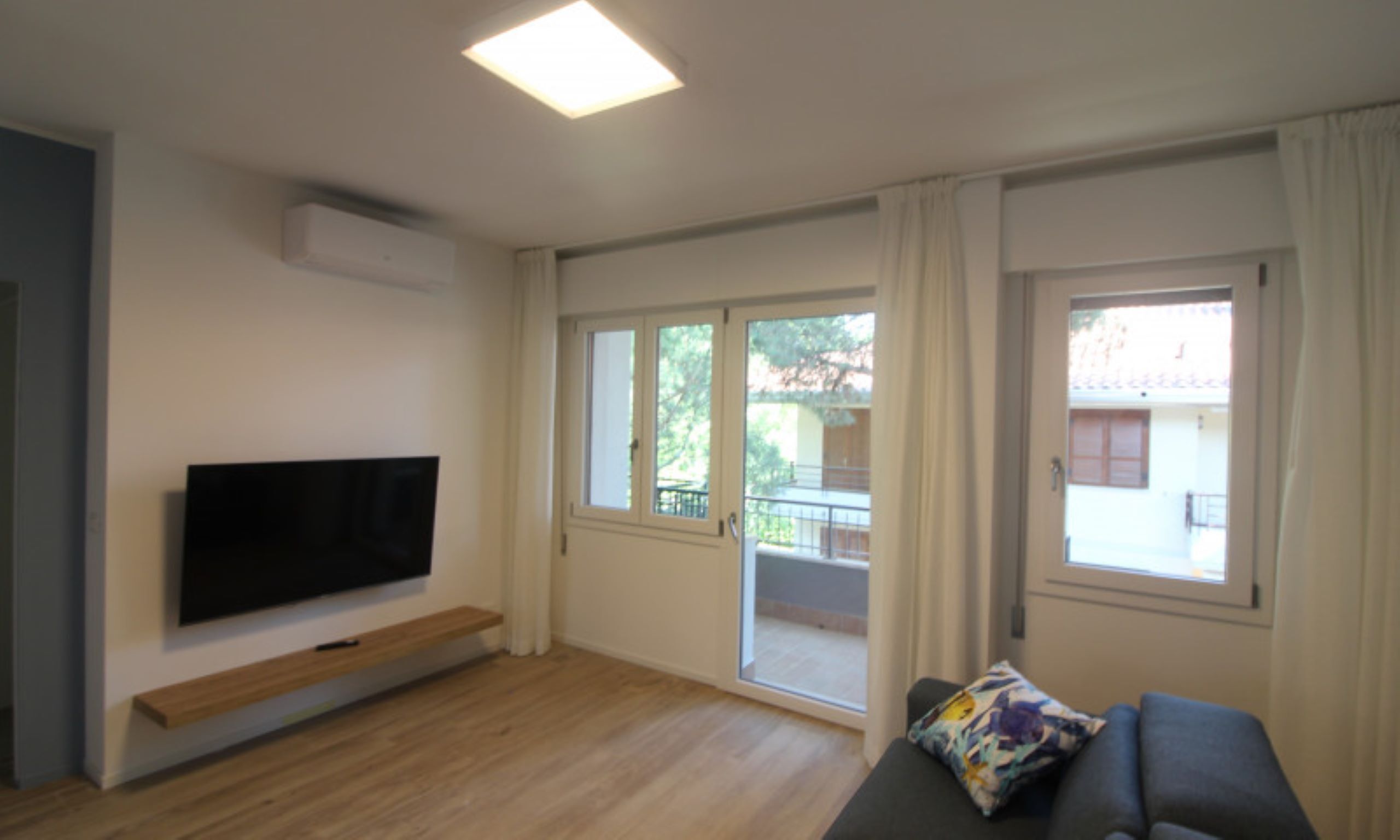Un ampio soggiorno con televisione a schermo piatto montata a parete, pavimento in legno, un divano grigio con cuscino decorato con motivi floreali e grandi finestre con tende.
