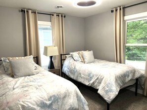 Et soveværelse med to senge, et natbord, en lampe og et vindue med gardiner.