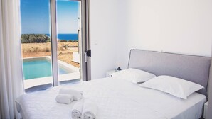 Ein Schlafzimmer mit einem Bett, zwei Kissen, einem Nachttisch, einer Lampe und Blick auf einen Pool und die Landschaft.