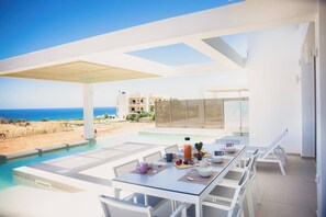 Un espace de restauration extérieur moderne, doté d’une piscine, de mobilier blanc et offrant une vue sur l’océan.