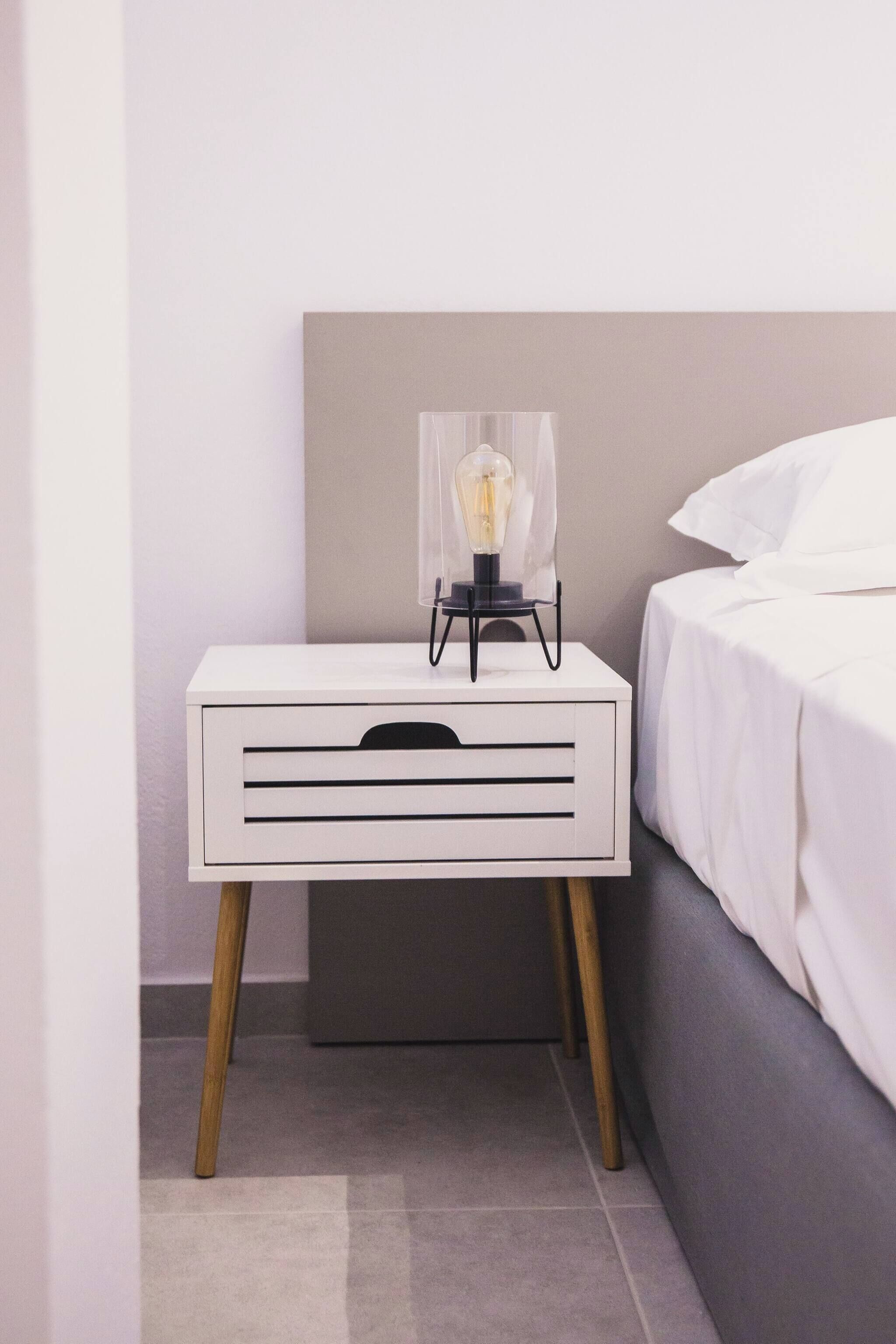 Una mesita de noche blanca con una lámpara, un cabecero gris y una cama gris.
