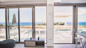 Ein modernes Wohnzimmer mit großen Schiebeglastüren, einem grauen Sitzsack, einem kleinen Tisch und Blick auf das Meer und den Poolbereich.