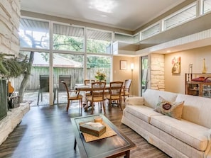 A spacious living room with a large window, a dining area with wooden furniture, and a cozy seating area with a sofa and cushions.