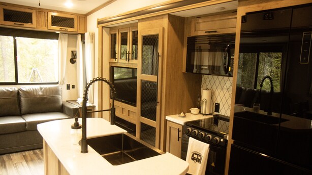 Een moderne camperkeuken met een centraal eiland, een ingebouwde oven en een groot raam met uitzicht op de natuur.