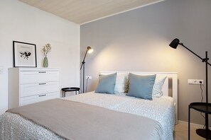 Un dormitorio con cama, mesitas de noche, cómoda, un cuadro enmarco y un florero con flores.