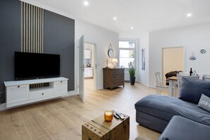 A modern living room with a flat-screen TV, a sofa, a wooden coffee table, and a dining area with a table and chairs.