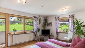 Un dormitorio con cama, televisión, sillón y ventanas amplias.