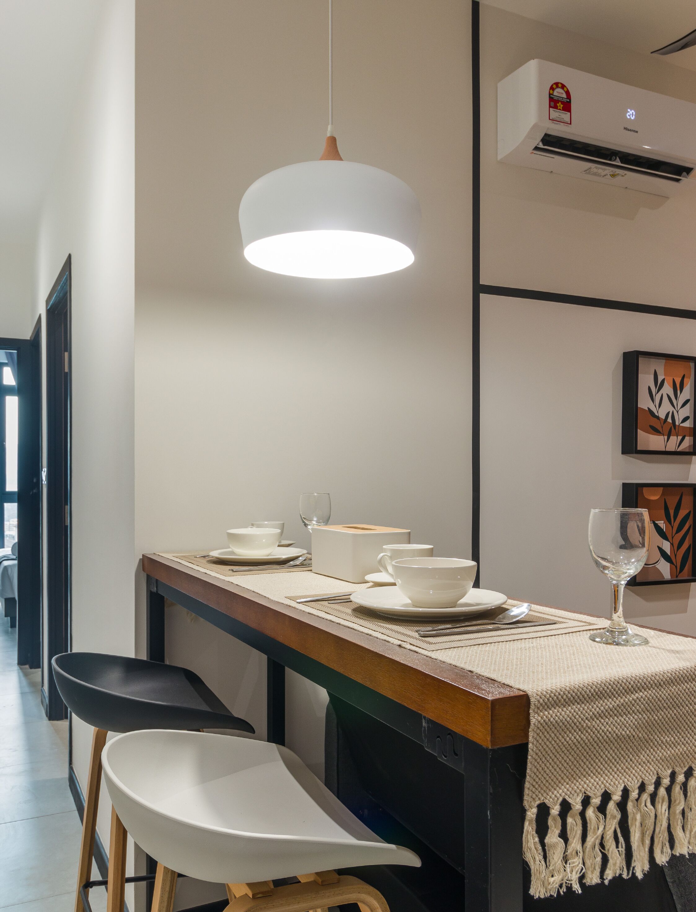 Uma área de refeições com uma mesa comprida posta para dois, uma luminária pendente e um ar-condicionado fixado na parede.