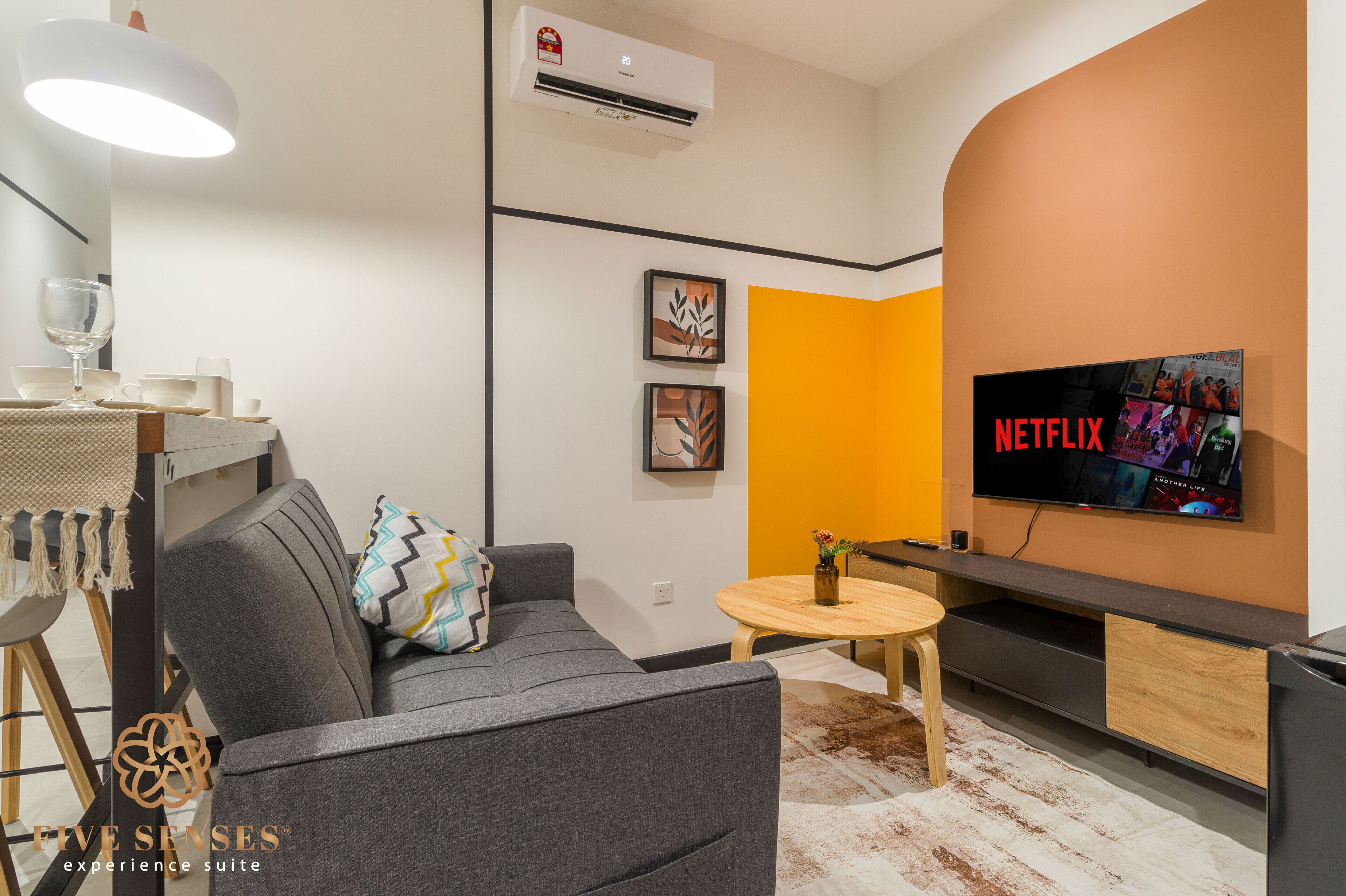 Un salon moderne avec un canapé gris, une petite table ronde, un téléviseur à écran plat fixé au mur et un appareil de climatisation.