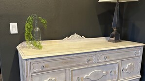 Un aparador de estilo antiguo con tiradores ornamentales y una superficie de cristal, situado frente a una pared oscura con una lámpara a su lado.