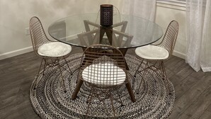Un comedor con una mesa de vidrio, sillas de alambre, un tapete estampado, una araña y un cuadro con flores en la pared.