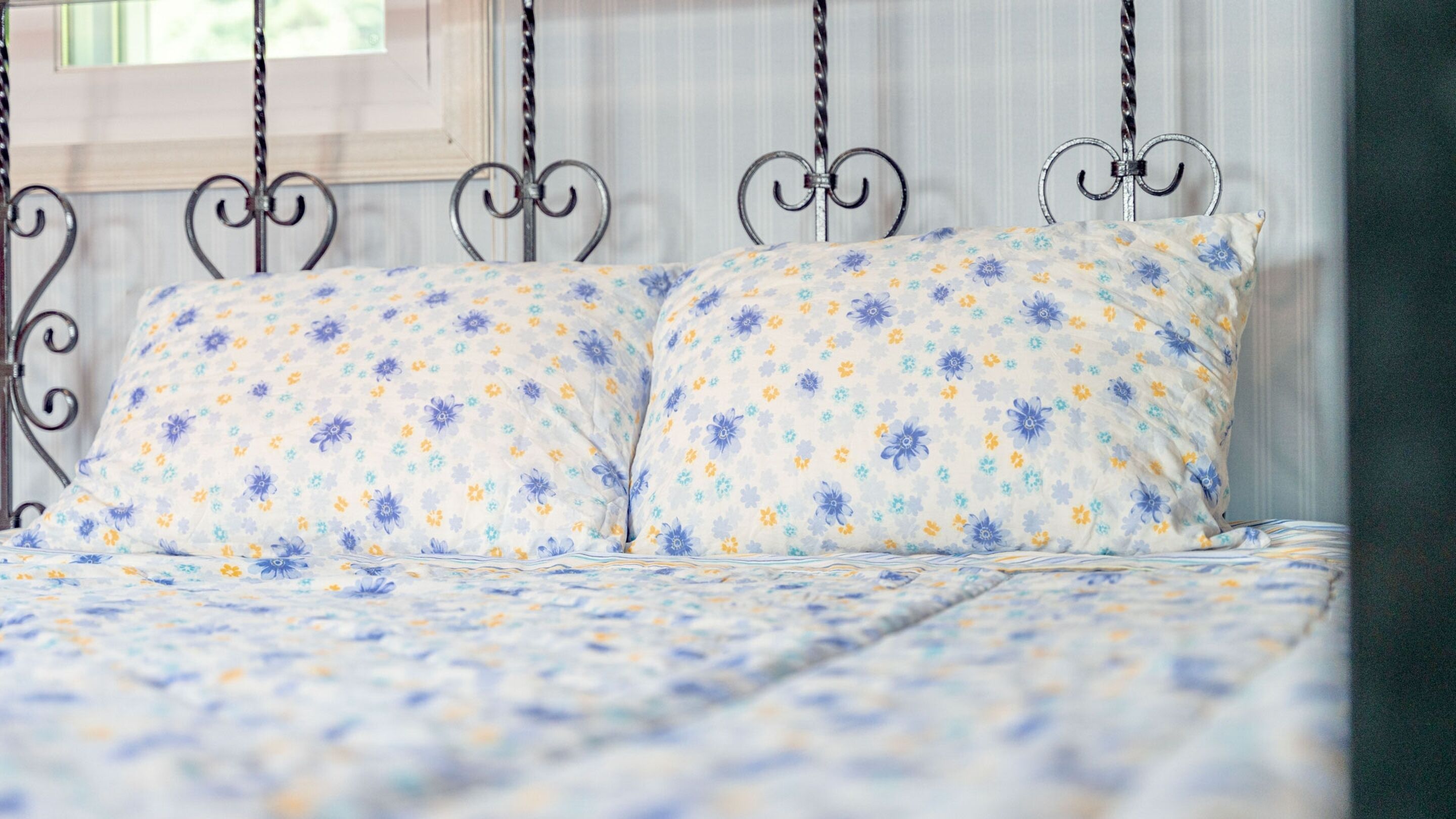 A bed with a floral-patterned bedspread and pillows, a metal headboard with a twisted design, and a window with blinds in the background.