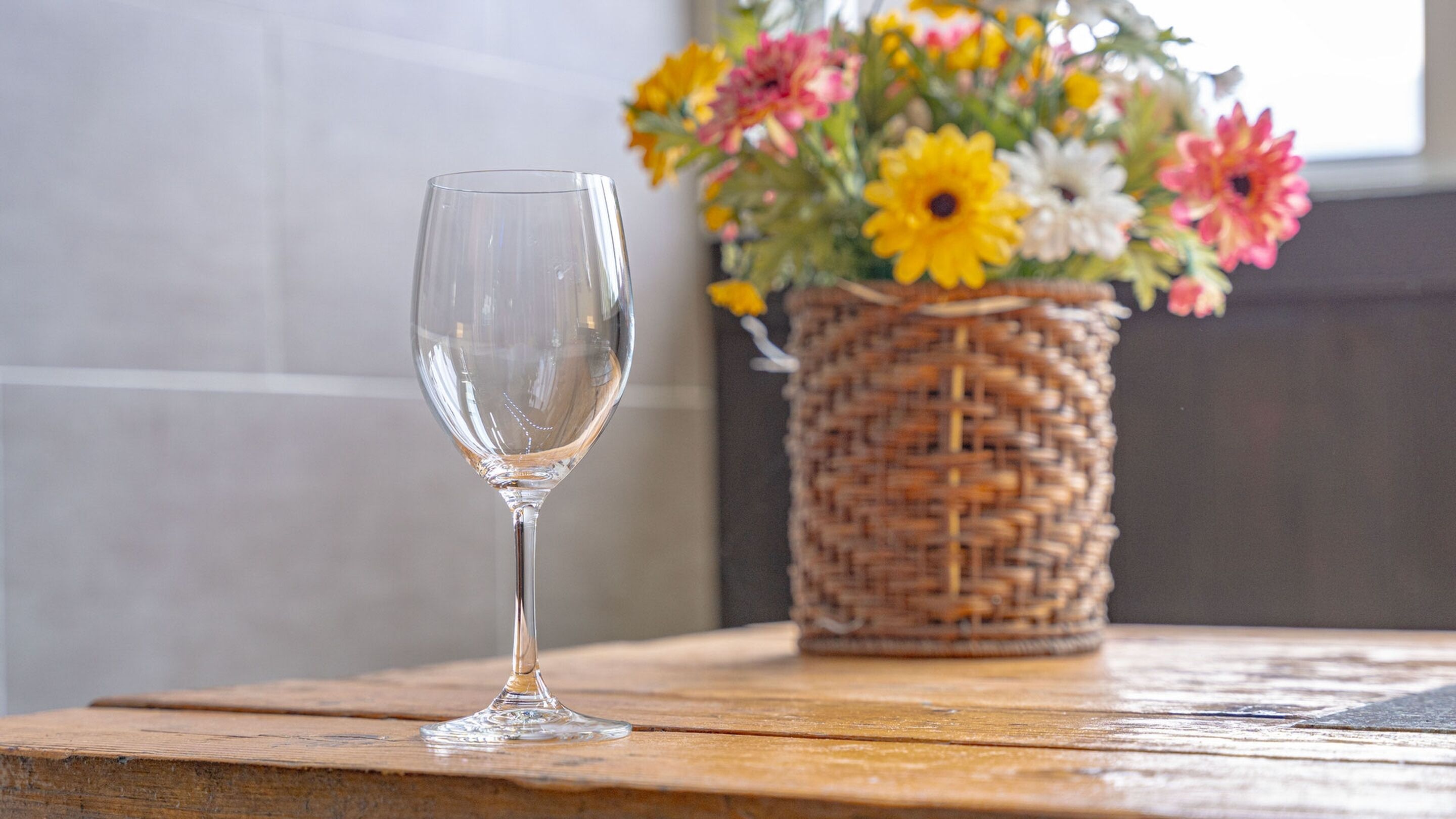 A clear glass on a wooden table with a wicker basket of flowers in the background.