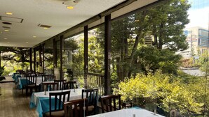 Un espace de restauration intérieur avec des tables et des chaises, de grandes fenêtres et une vue sur les arbres et les bâtiments à l’extérieur.