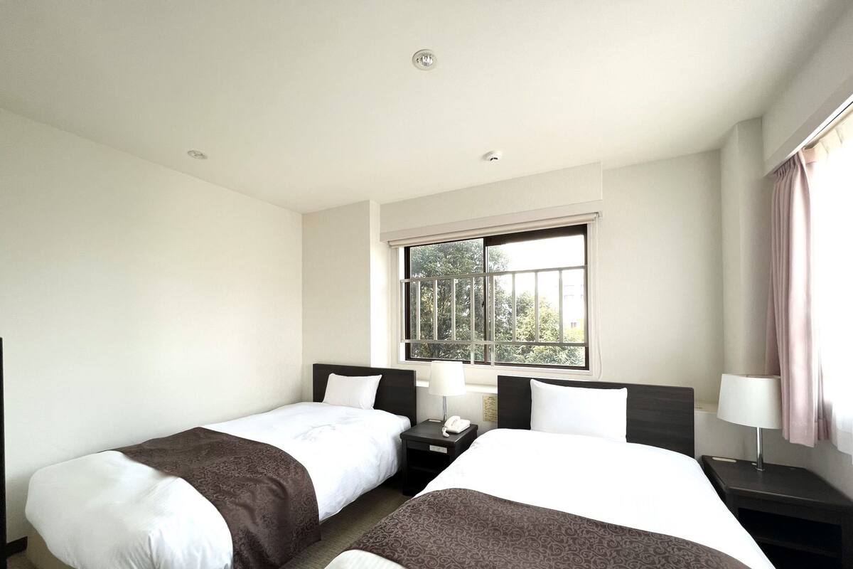 A hotel room with two beds, a window with a view of trees, and a nightstand with a lamp.