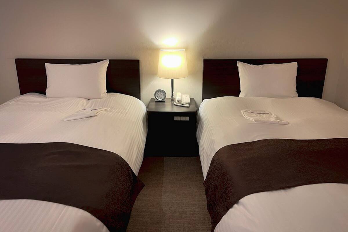 A hotel room with two single beds, a bedside table with a lamp, a clock, and a phone.