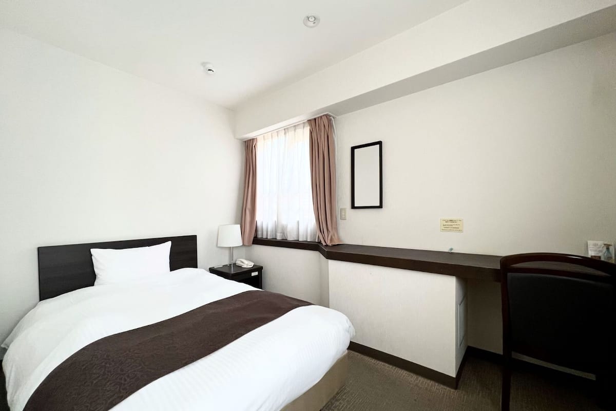 A hotel room with a bed, a desk, a chair, a nightstand, a lamp, a window with curtains, and a framed picture on the wall.