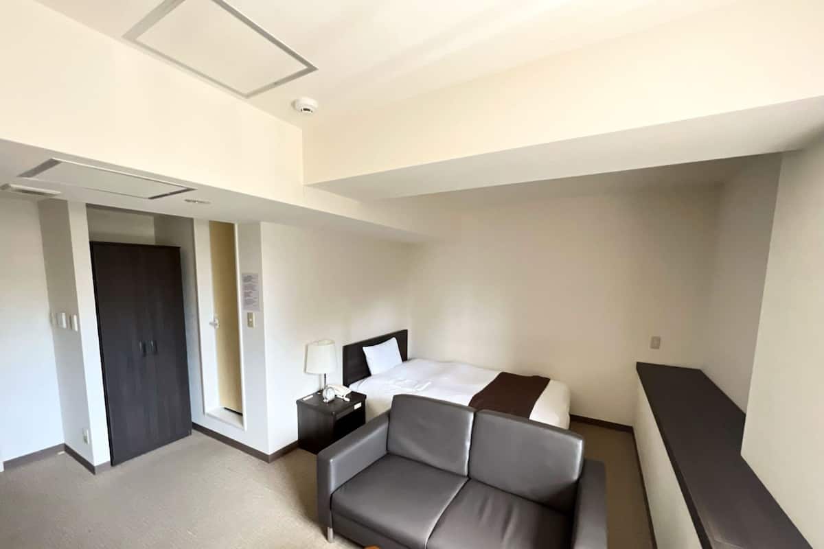 A hotel room with a bed, a sofa, a nightstand with a lamp, a desk, and a door.