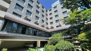 Un hôtel à plusieurs étages au design moderne, caractérisé par une façade combinant verre et pierre, un espace de stationnement et un jardin bien entretenu agrémenté d’une lanterne en pierre traditionnelle.
