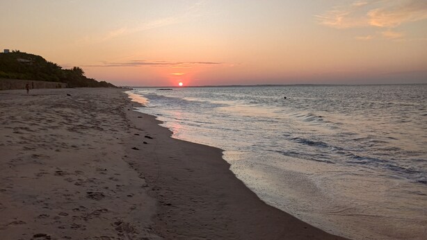 A beach with footprints leading to the water, a sunset in the background, and a distant shoreline. - Casa em Paracuru Beach (Paracuru)