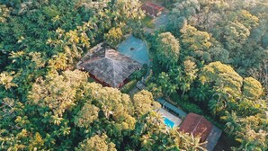 Vista aérea de uma propriedade com um edifício central rodeado por vegetação tropical densa, uma piscina e uma construção adjacente.