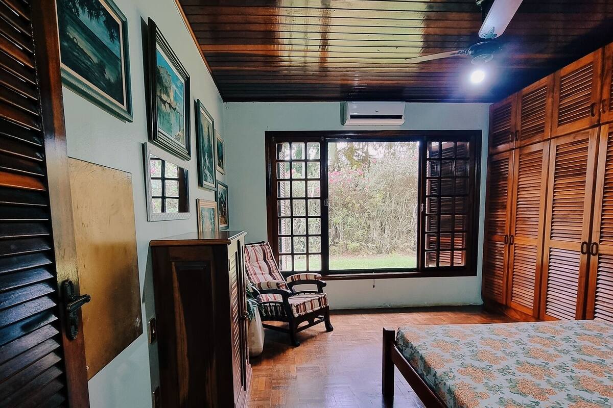 A room with wooden flooring, a bed with a floral bedspread, a chair, a wooden wardrobe, and a door with a window.