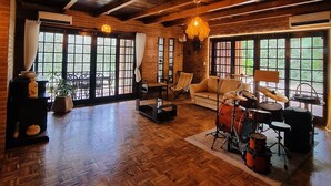 Um quarto espaçoso com piso de madeira, janelas amplas, um sofá, um set de bateria e uma lareira.