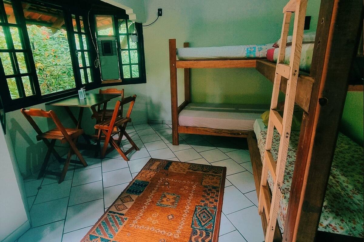 A room with a bunk bed, a small table with chairs, a window with blinds, and a patterned rug on the floor.