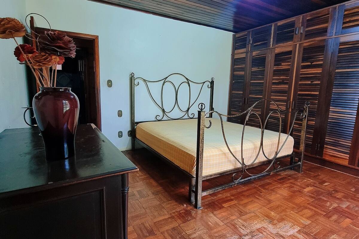A bedroom with a wooden floor, a metal bed frame, a wooden dresser, and a vase with dried flowers.