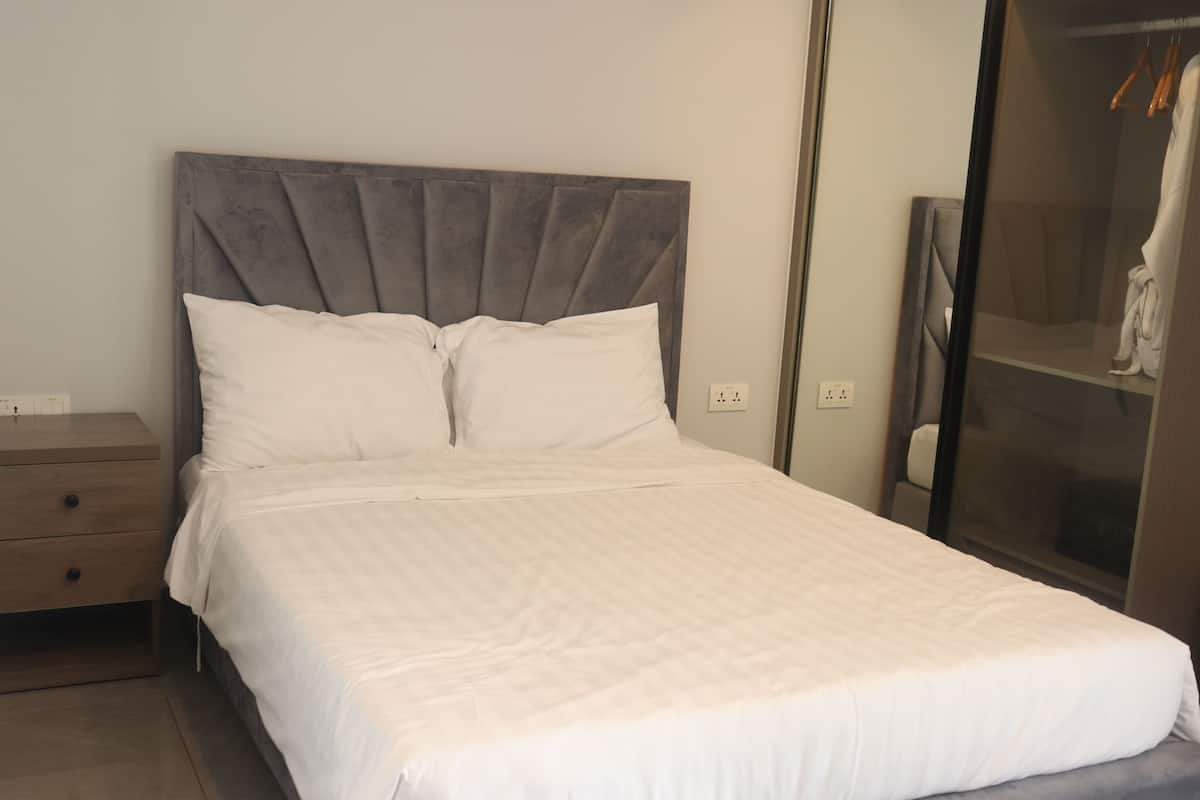 Ein ordentlich gemachtes Bett mit grauem Stoffkopfteil, einem Holfnachtkästchen und einem begehbaren Kleiderschrank mit Kleidung.
