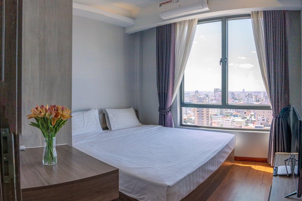 Ein Hotelzimmer mit einem großen Bett, einem Nachttisch mit Blumenvase, einem Schreibtisch mit Computer und Blick auf die Stadt durch ein großes Fenster.