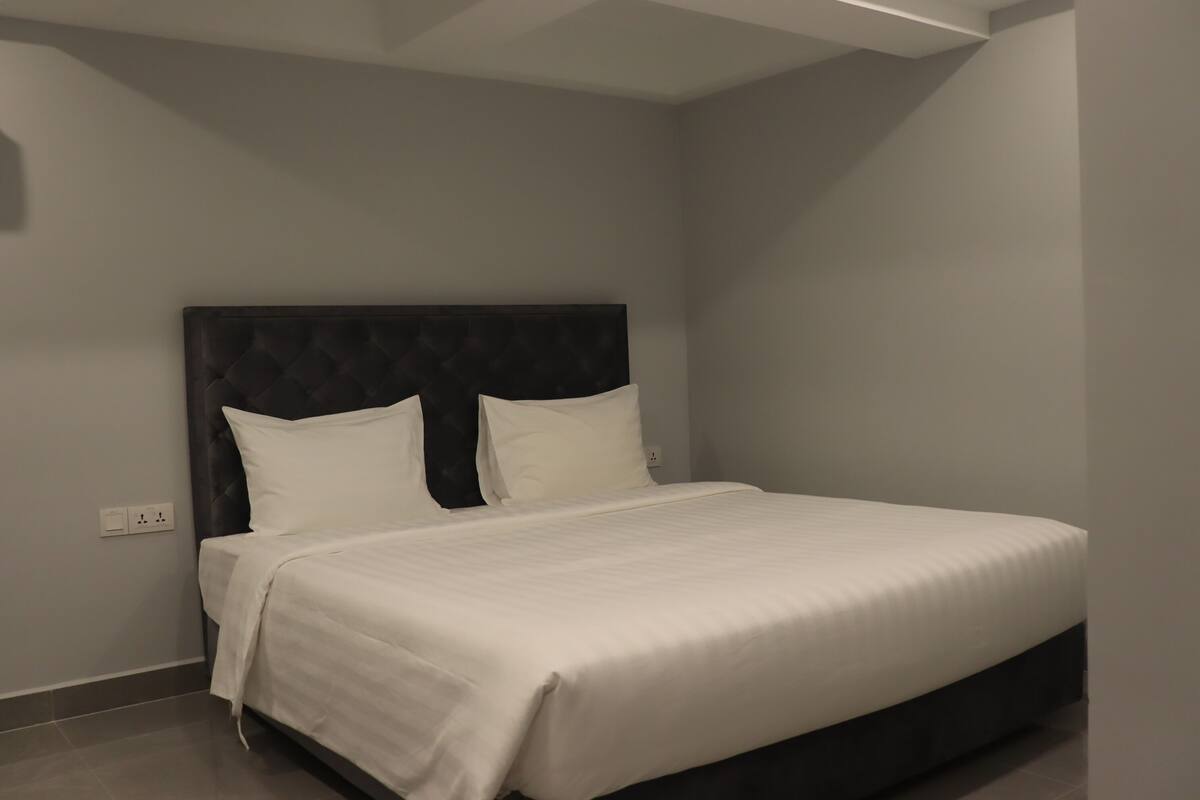 Ein ordentlich bezogenes Bett mit dunklem Kopfteil und weißer Bettwäsche in einem modernen Zimmer mit schlichter Wand und sichtbarer Steckdose.