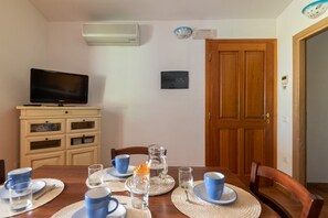 A dining area with a table set for four, a television on a cabinet, and a wooden door in the background. - Stunning Residence Bouganvillage 2 Bedroom Sleeps 6 Child (Budoni)