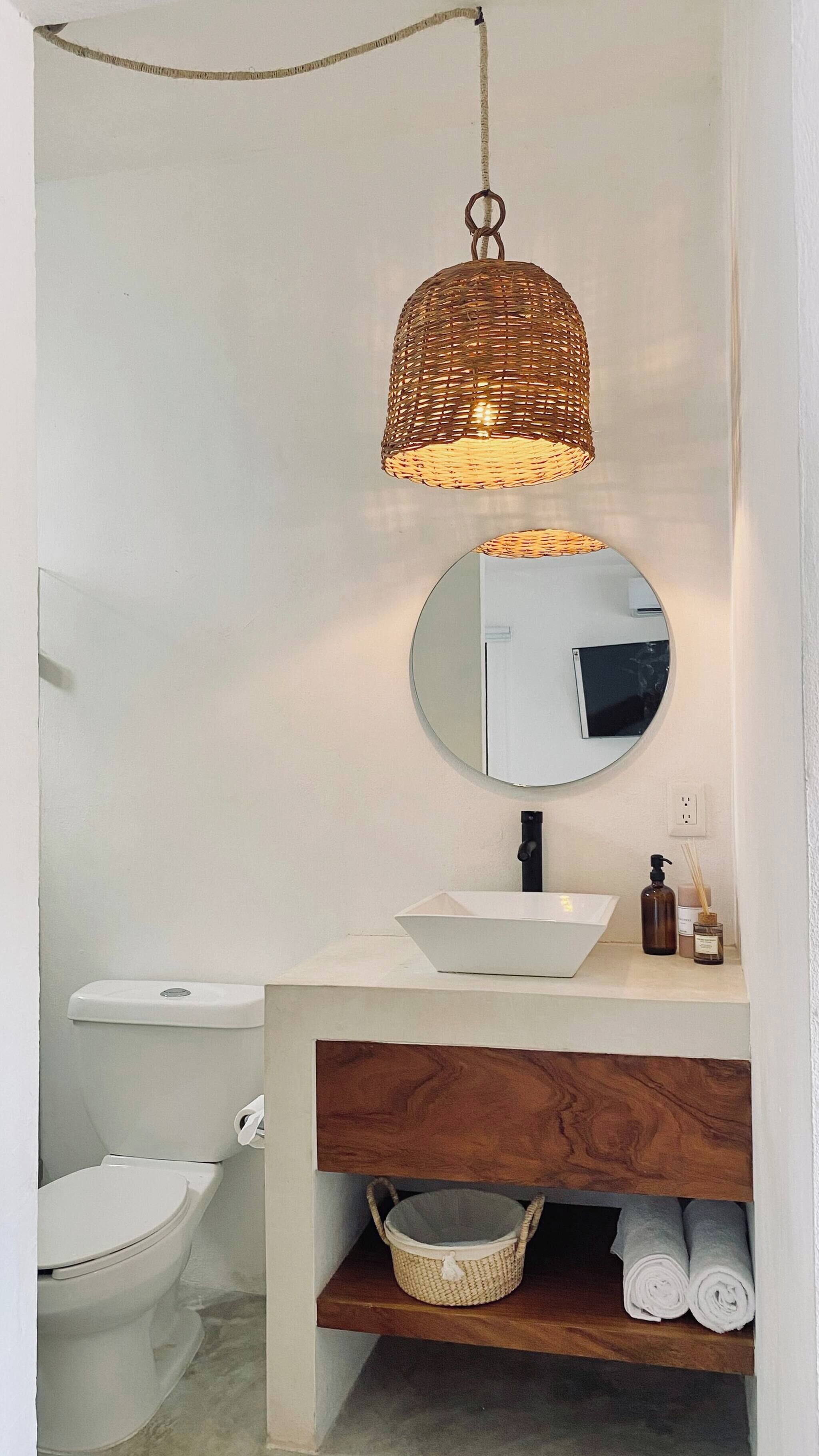 Een badkamer met een wit toilet, een ronde spiegel, een witte wastafel, een houten wastafelmeubel, een hanglamp van wilg en een mandje van wilg op het meubel.