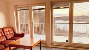 A room with a brown leather sofa, a wooden table, and large sliding glass doors leading to a balcony with a view of a body of water and trees. - Bjurvik's Villa - Flat 2 - Business Customers Only (Flen)