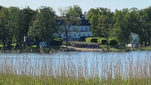 Un grand bâtiment blanc à la façade symétrique, entouré d’une végétation luxuriante et bordé d’une étendue d’eau calme.