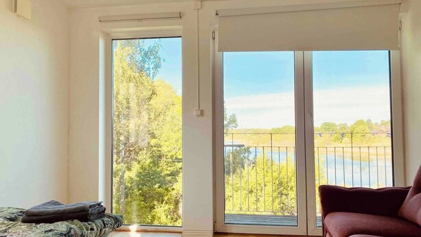 Une pièce dotée d’une grande porte-fenêtre coulissante qui donne sur un balcon offrant une vue sur des arbres et un plan d’eau. On y trouve un canapé à motifs et un fauteuil rouge.