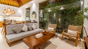 A modern living room with a wooden ceiling, a sofa with cushions, a coffee table, wicker chairs, and a large window with bamboo plants. - Top Choice 3 Bedrooms Pool Villa in Central Canggu (Canggu)