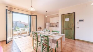 A dining area with a table set for four, green chairs, a sliding glass door leading to a balcony, and a kitchen area with white cabinets and a wooden door.