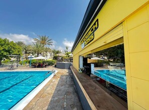 Un espace piscine avec un bâtiment jaune portant l’inscription « Sandwich Inn » et un ciel d’un bleu limpide.