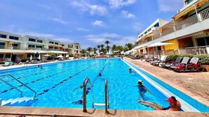 Ein Schwimmbecken mit klarem, blauem Wasser, umgeben von Liegest ühlen und Sonnenschirmen, mit Gebäuden im Hintergrund.