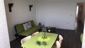 Un coin repas avec une table, des chaises et une nappe verte. On y trouve un canapé avec un coussin vert et un objet décoratif accroché au mur.