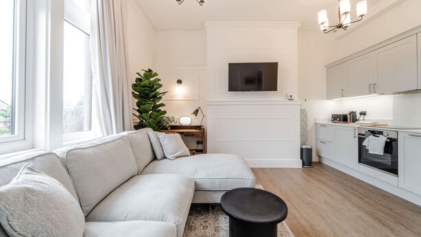 A modern living room with a sectional sofa, a round black coffee table, a flat-screen TV mounted on the wall, and a kitchen area with white cabinets and a built-in oven. - Luxury Apartment In The Heart Of Hoylake (Wirral)