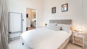 A neatly arranged bedroom with a large bed, bedside tables, a nightstand, a mirror, and a door leading to a bathroom. - Luxury Apartment In The Heart Of Hoylake (Wirral)