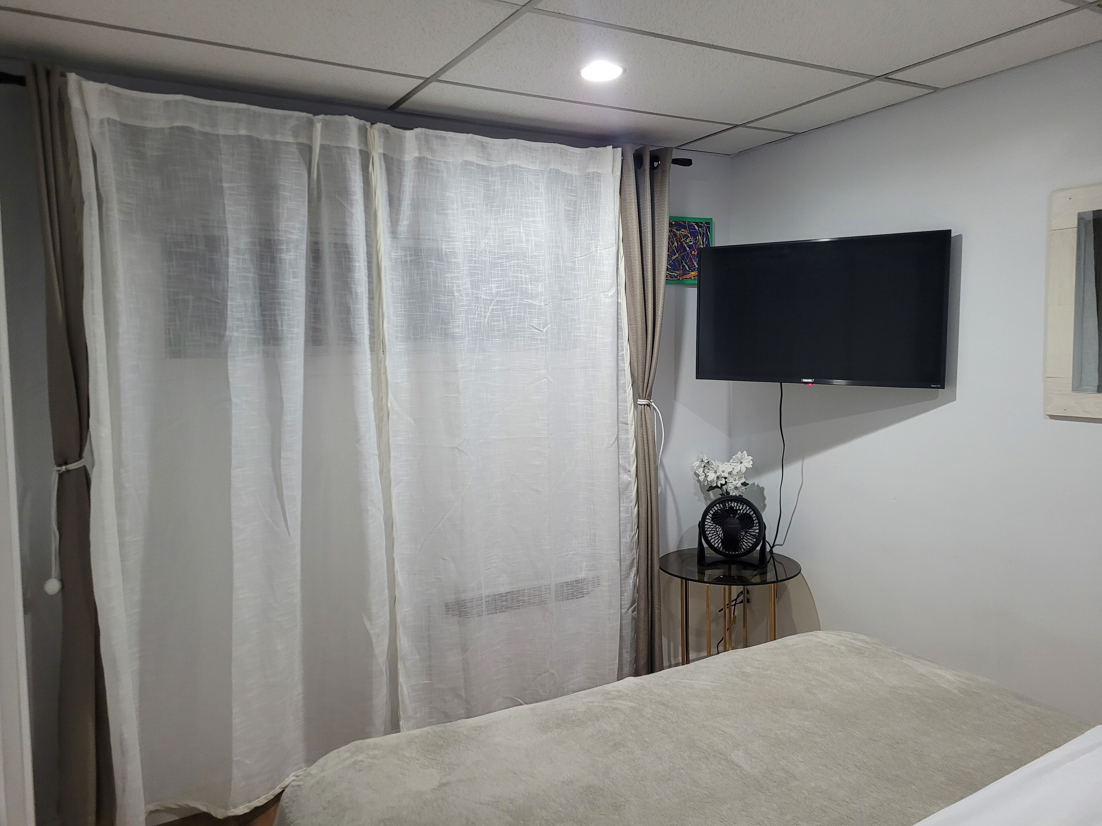 A hotel room with a bed, a TV mounted on the wall, a small table with a fan, and sheer curtains.
